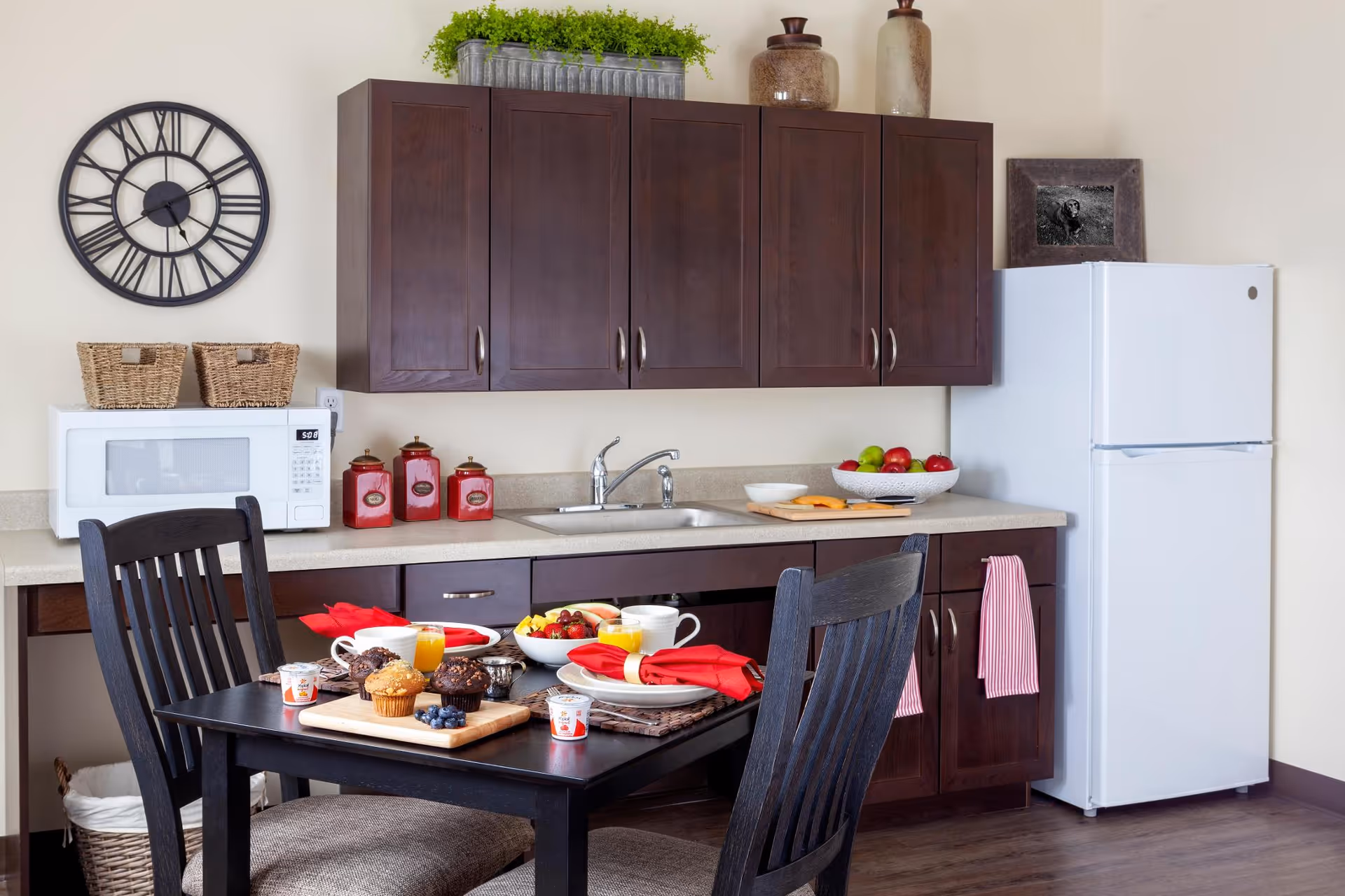 A small kitchen area with dark wooden cabinets, a white microwave, a white refrigerator, and a countertop with a sink. On the countertop, there are decorative jars, a bowl of fruit, and a cutting board with cheese. In front of the kitchen is a black dining table set with plates, cups, muffins, fruit, and orange juice, along with red napkins. A large black wall clock is mounted on the wall above the microwave.