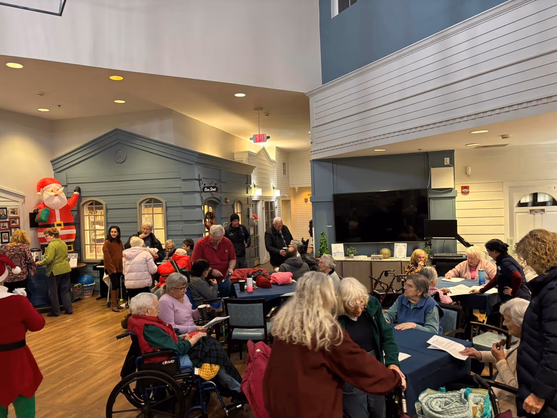 A lively indoor common area in a senior living facility with many elderly residents and caregivers engaged in various activities. The room features a large TV mounted on the wall, tables with chairs, and a decorative blue house-like structure. There is a large inflatable Santa Claus decoration near the back wall, indicating a festive holiday atmosphere.
