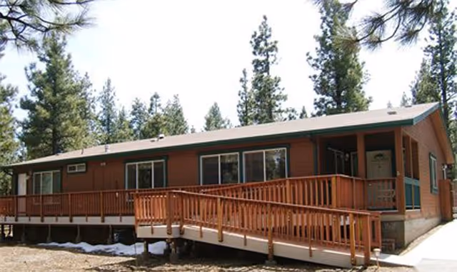 Single-story brown building with a wooden ramp and railing leading to the entrance, surrounded by tall pine trees under a clear sky.