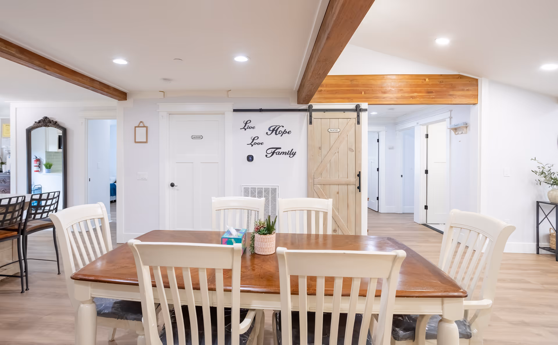 A bright dining area with a wooden table and six white chairs. The room features light wood flooring, white walls, and exposed wooden beams on the ceiling. On the wall, there is a decorative sign with the words 'Live Hope Love Family'. A sliding barn door labeled 'Pantry' is visible, along with several white doors leading to other rooms. A small plant and a tissue box are placed on the dining table.