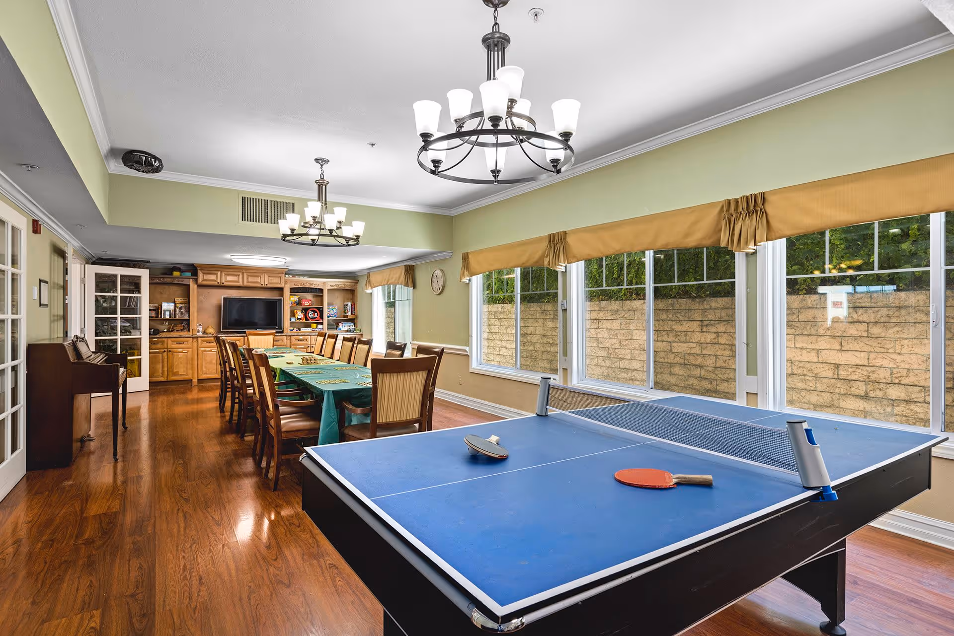 A spacious recreational room with a blue ping pong table in the foreground, two paddles and a ball on it. Behind the table is a long table covered with a green tablecloth surrounded by wooden chairs. The room has large windows with yellow valances, wooden flooring, two chandeliers, and a built-in wooden cabinet with a TV and board games.