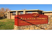 Exterior view of a single-story building with stone and brick facade under a clear blue sky. In front of the building is a large red sign with gold lettering that reads 'Atlantic Shores Rehabilitation & Health Center'. There is green grass and a tree with autumn-colored leaves on the left side.