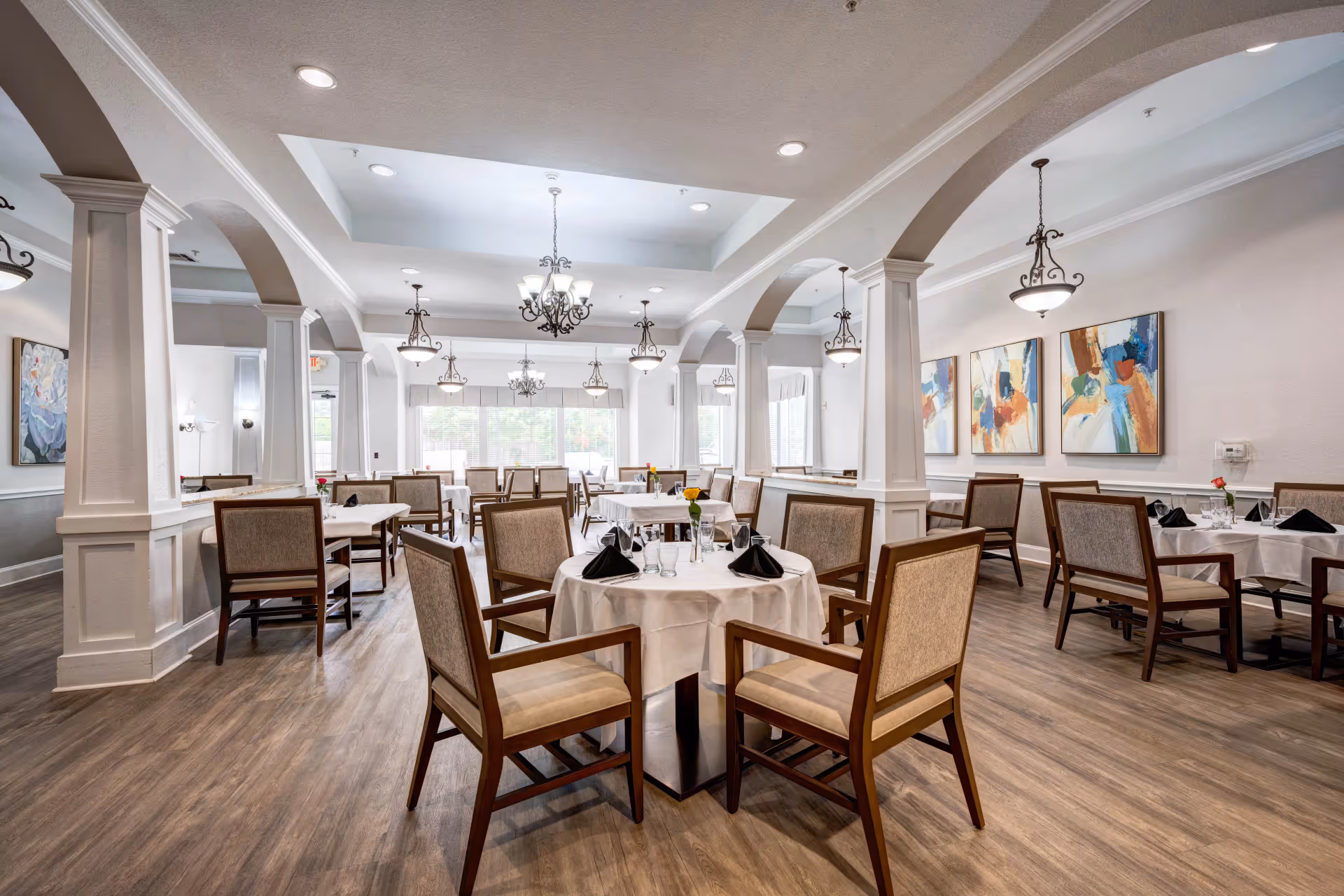 A spacious and elegant dining room with multiple round tables covered with white tablecloths, each set with black napkins, glasses, and small flower vases. The room features wooden chairs with cushioned seats and backs, large windows allowing natural light, decorative ceiling lights, and colorful abstract paintings on the walls.