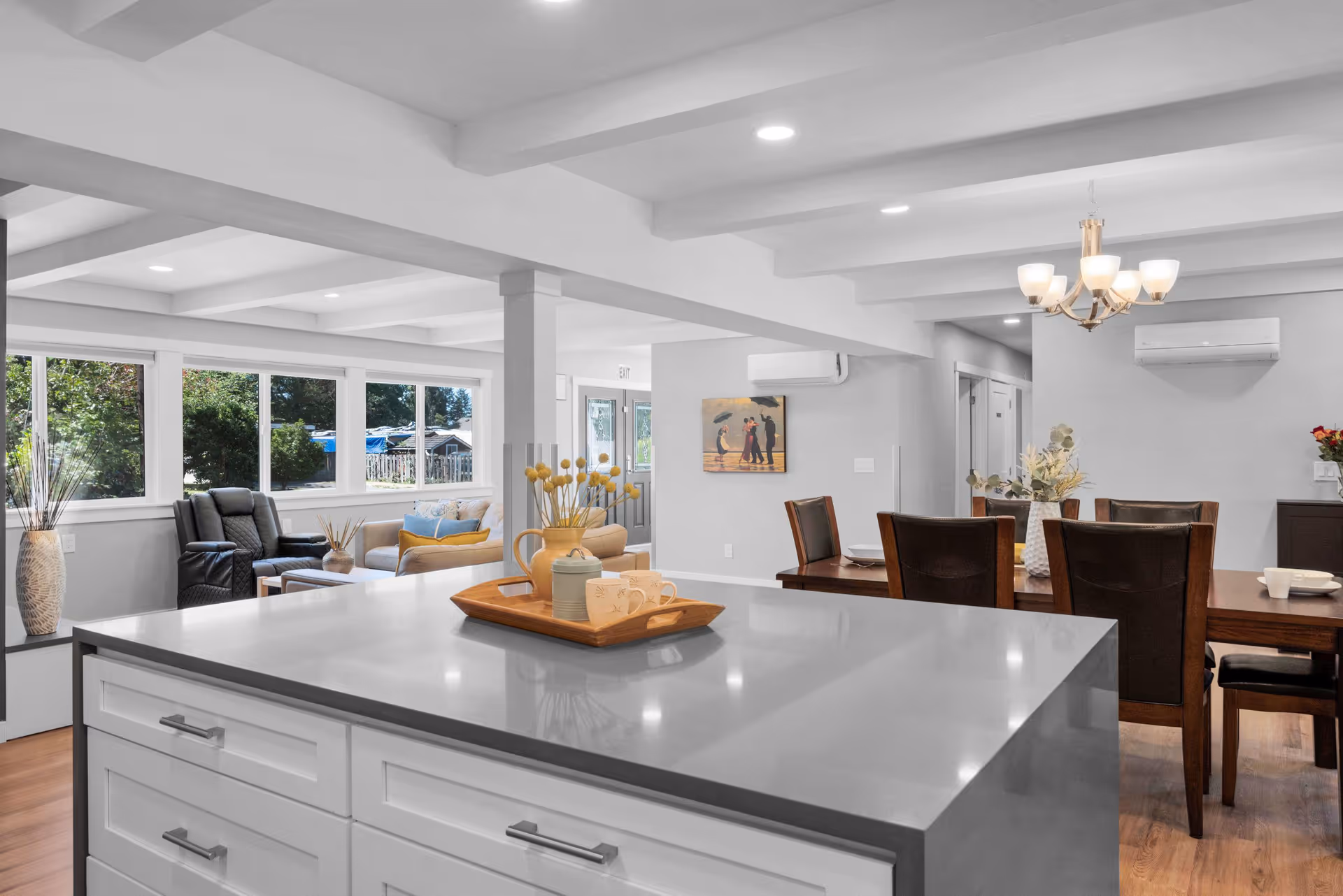 Bright and modern open-concept living space featuring a large kitchen island with a tray holding a teapot and cups, a dining area with a wooden table and six chairs under a chandelier, and a cozy sitting area with a black recliner and beige sofa near large windows showing greenery outside.
