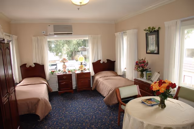 A bright and cozy bedroom with two single beds covered in beige bedspreads. Between the beds are two wooden nightstands with decorative lamps and small plants. The room has large windows with white curtains letting in natural light. There is a round table with a white tablecloth and a vase of colorful flowers, surrounded by chairs. The walls are light-colored, and there is a framed picture and a bouquet of flowers on a dresser. The floor is covered with a blue patterned carpet.