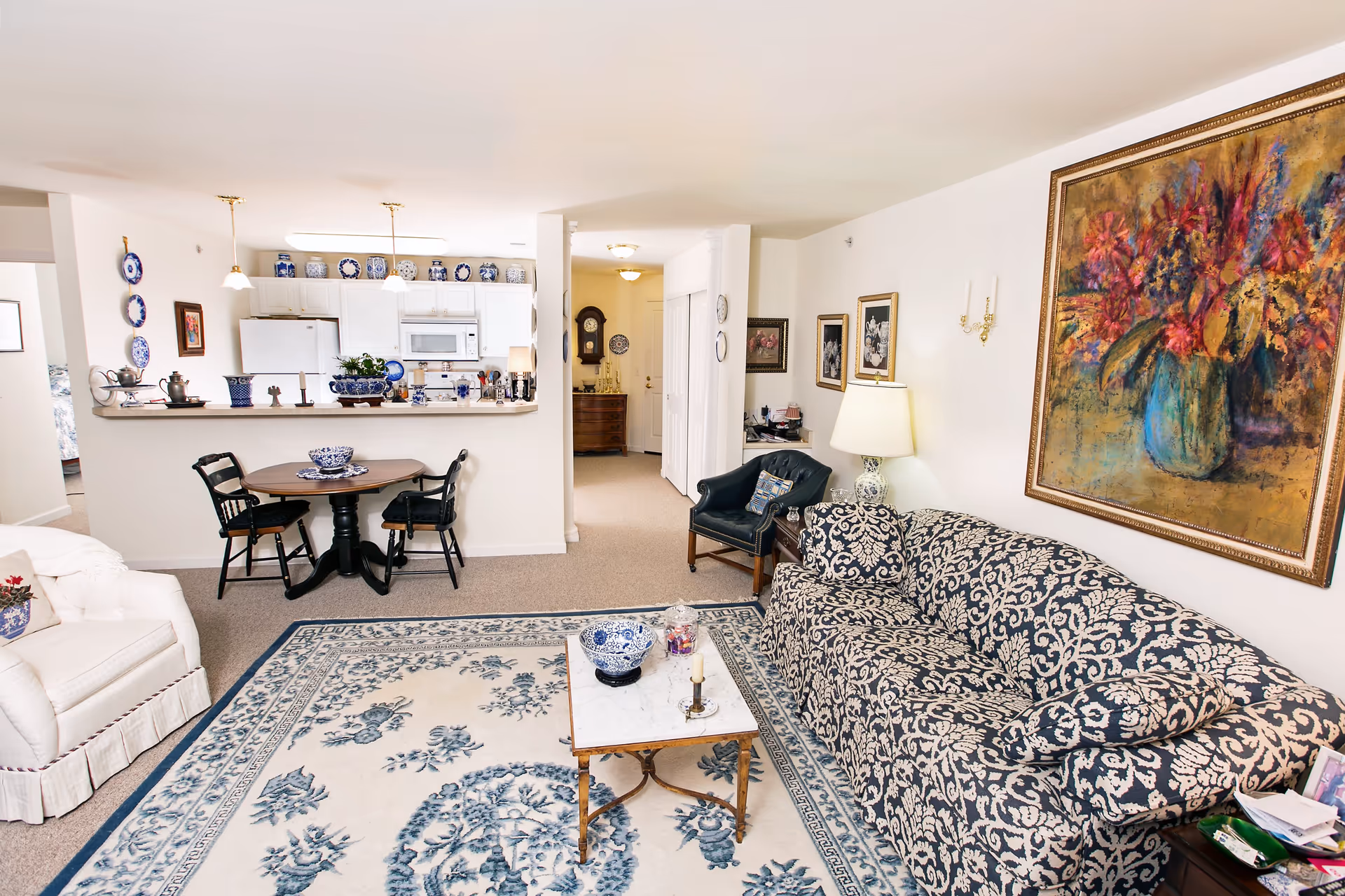 A cozy living room and kitchen area in a senior living community. The living room features a patterned blue and white sofa, a white armchair, a small marble-top coffee table with decorative items, and a large floral painting on the wall. The kitchen has white cabinets, a refrigerator, a microwave, and a small round dining table with two chairs. Blue and white decorative plates and vases are displayed on shelves and walls. The space is well-lit with ceiling lights and a table lamp.