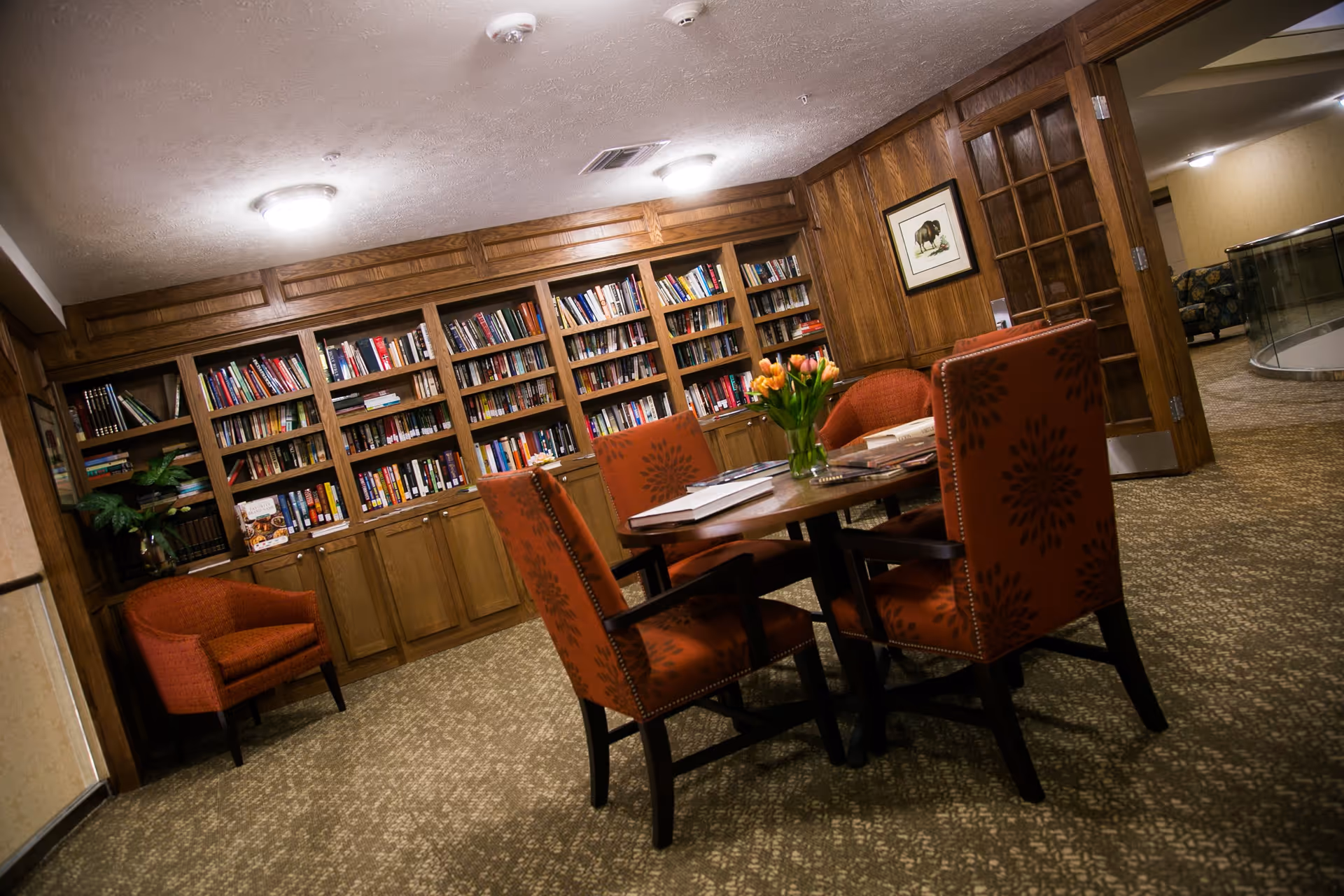 A cozy reading room with wooden bookshelves filled with books along the wall, a round table with four orange patterned chairs around it, and a vase of orange tulips on the table. There is a framed picture on the wood-paneled wall and an orange armchair in the corner. The room has carpeted flooring and warm lighting.