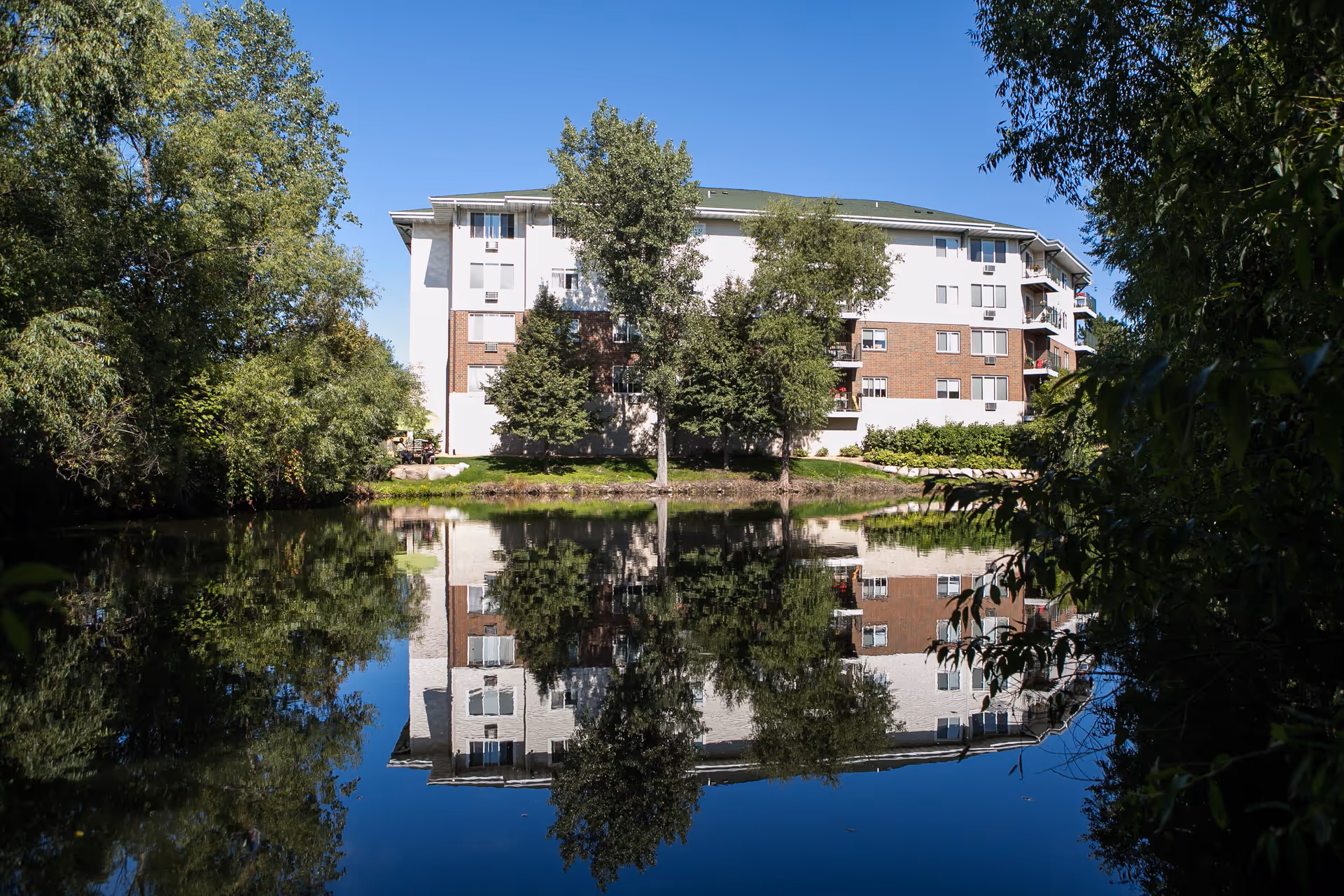 A multi-story retirement community building with white and brick exterior walls, surrounded by trees and greenery, reflected clearly in a calm pond in front of it under a clear blue sky.