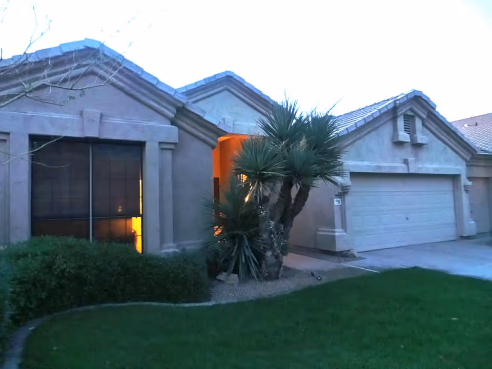 Exterior view of a single-story residential building with a two-car garage, a large window with blinds, and a small landscaped area with green grass, bushes, and a palm-like tree near the entrance. The building has a stucco finish and a tiled roof, with warm light glowing from inside near the entrance.
