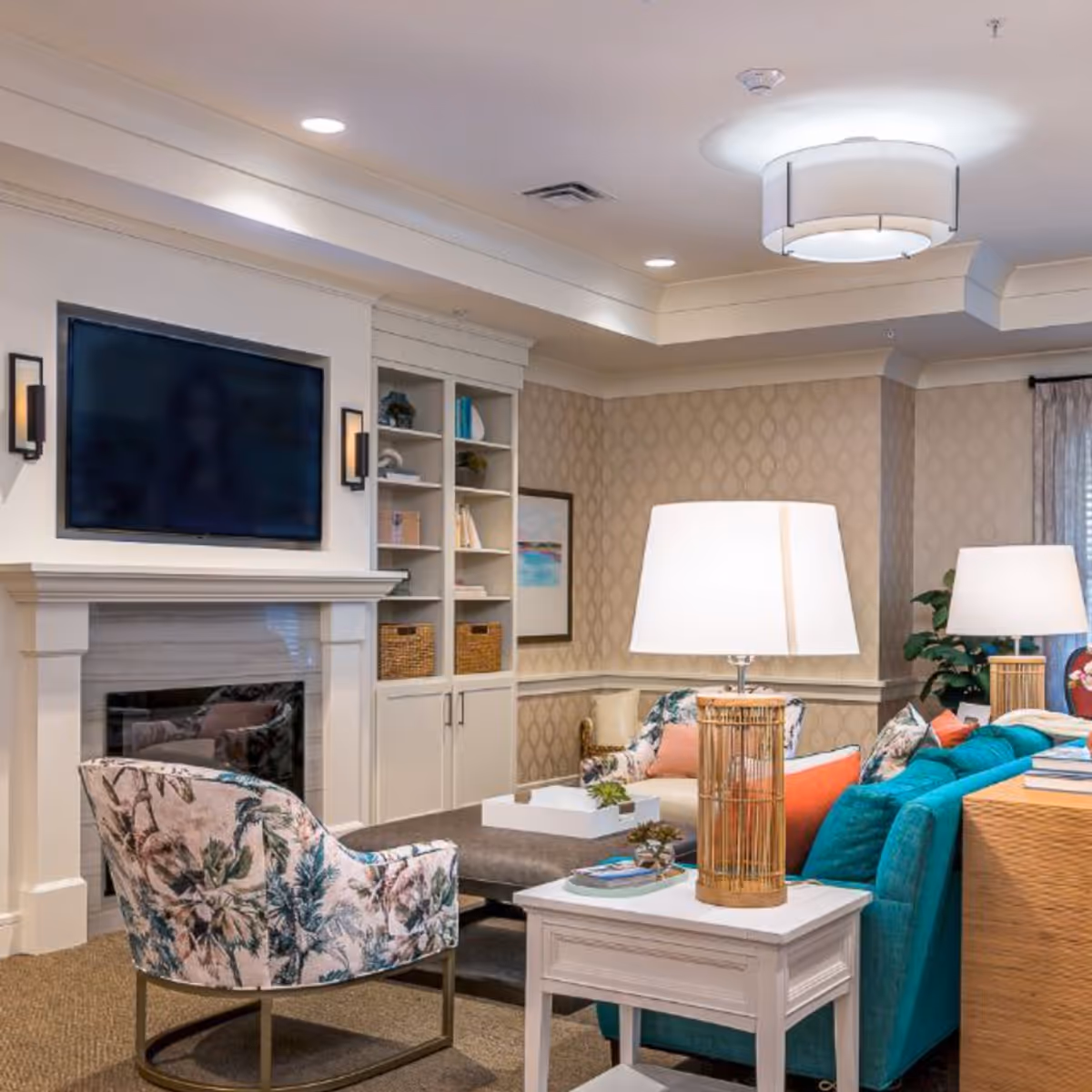 Bright, well-appointed common living room with a teal sofa, floral armchair, side tables with lamps, built-in shelving and a wall-mounted TV above a fireplace.