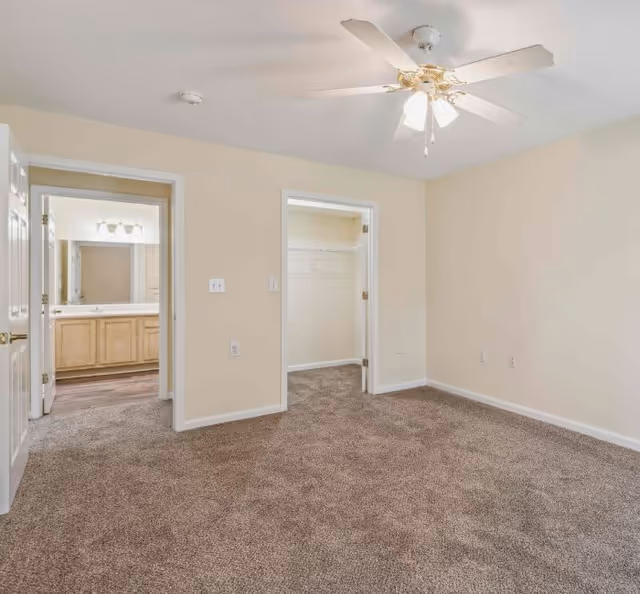 Empty bedroom with beige walls, brown carpet, a white ceiling fan with lights, and two doorways leading to a bathroom with a vanity and a walk-in closet.