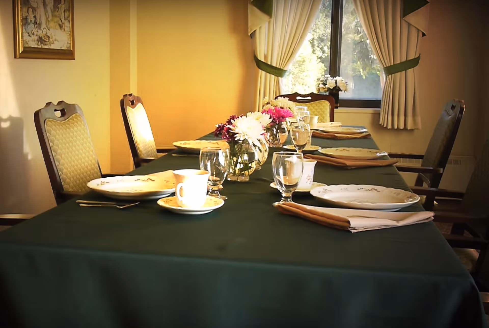 A dining table set with plates, cups, glasses, and napkins, decorated with small vases of flowers. The table is covered with a dark green tablecloth and surrounded by upholstered chairs. A window with curtains is visible in the background, letting in natural light.