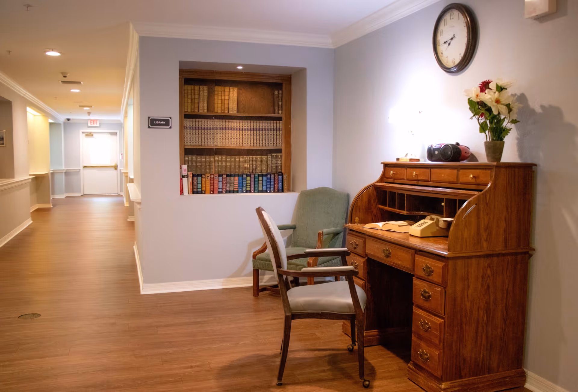 A hallway in a senior living facility with wooden flooring and light-colored walls. On the right side, there is a wooden roll-top desk with an old-fashioned telephone, a radio, and a vase with flowers on top. Two chairs, one green and one with a light cushion, are placed near the desk. A clock hangs on the wall above the desk. A built-in bookshelf labeled 'Library' is inset into the wall, filled with books. The hallway extends toward a door with an exit sign above it.