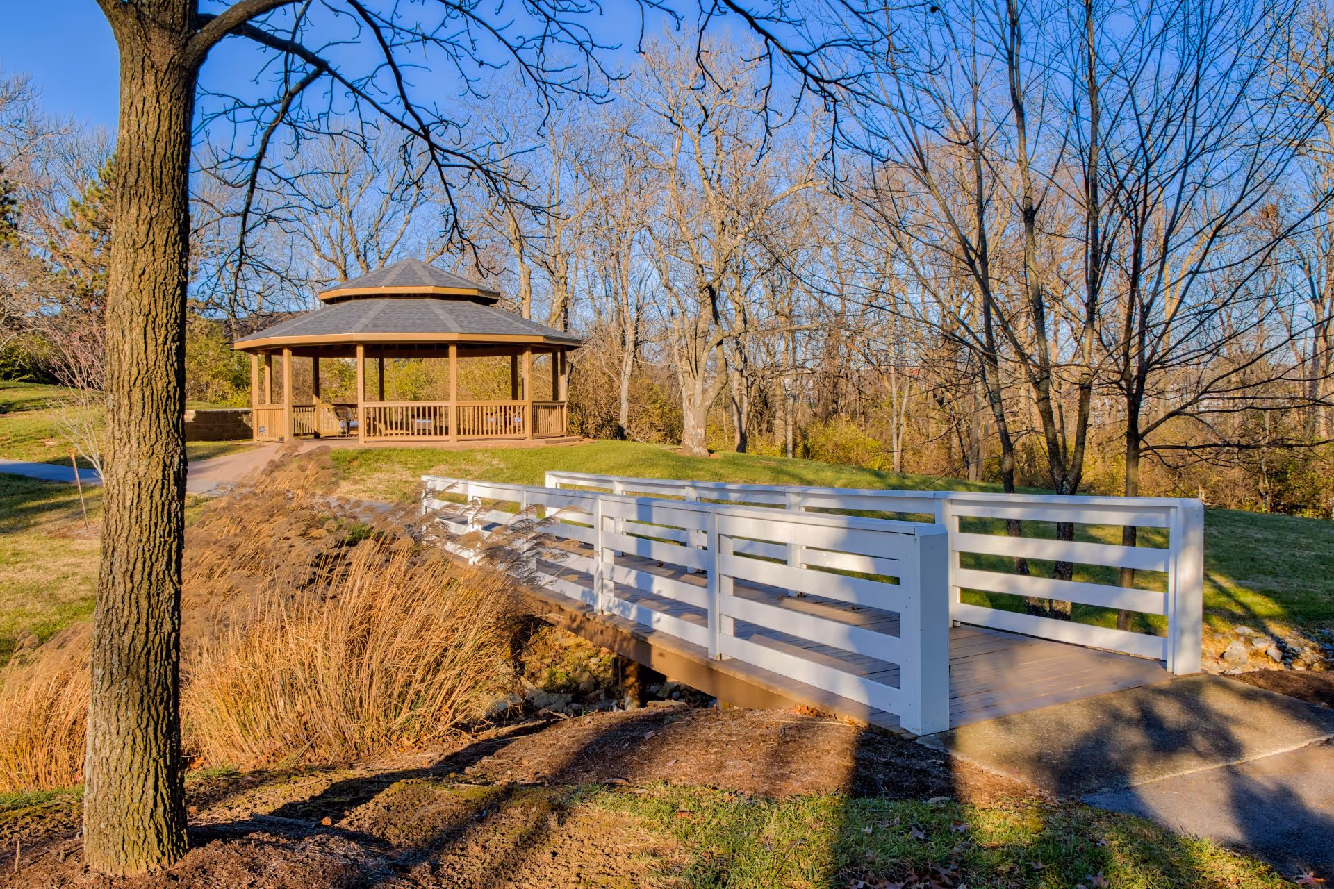 A white wooden bridge leading to a gazebo in a park-like outdoor setting with leafless trees and dry grass under a clear blue sky.