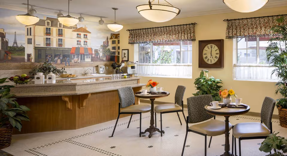 A cozy dining area in a senior living facility with two small round tables set with cups, saucers, and flower vases. The room features a countertop with a sink, coffee maker, and decorative items. Large windows with floral valances let in natural light, and a large wall clock is mounted between the windows. The walls have a mural depicting a European street scene with buildings and people. Potted plants add greenery to the space.