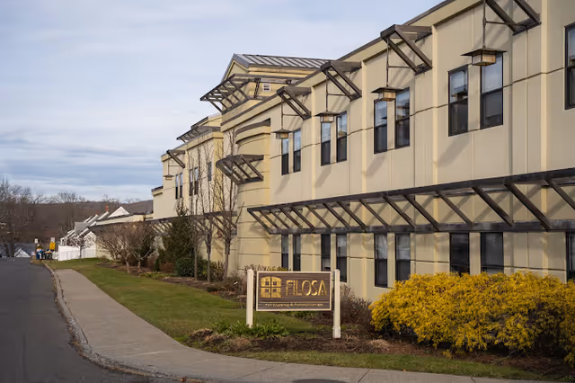 Exterior view of a beige multi-story building with multiple windows and metal awnings. A sidewalk runs alongside the building, with landscaped bushes and grass. A sign in front reads 'FILOSA'.