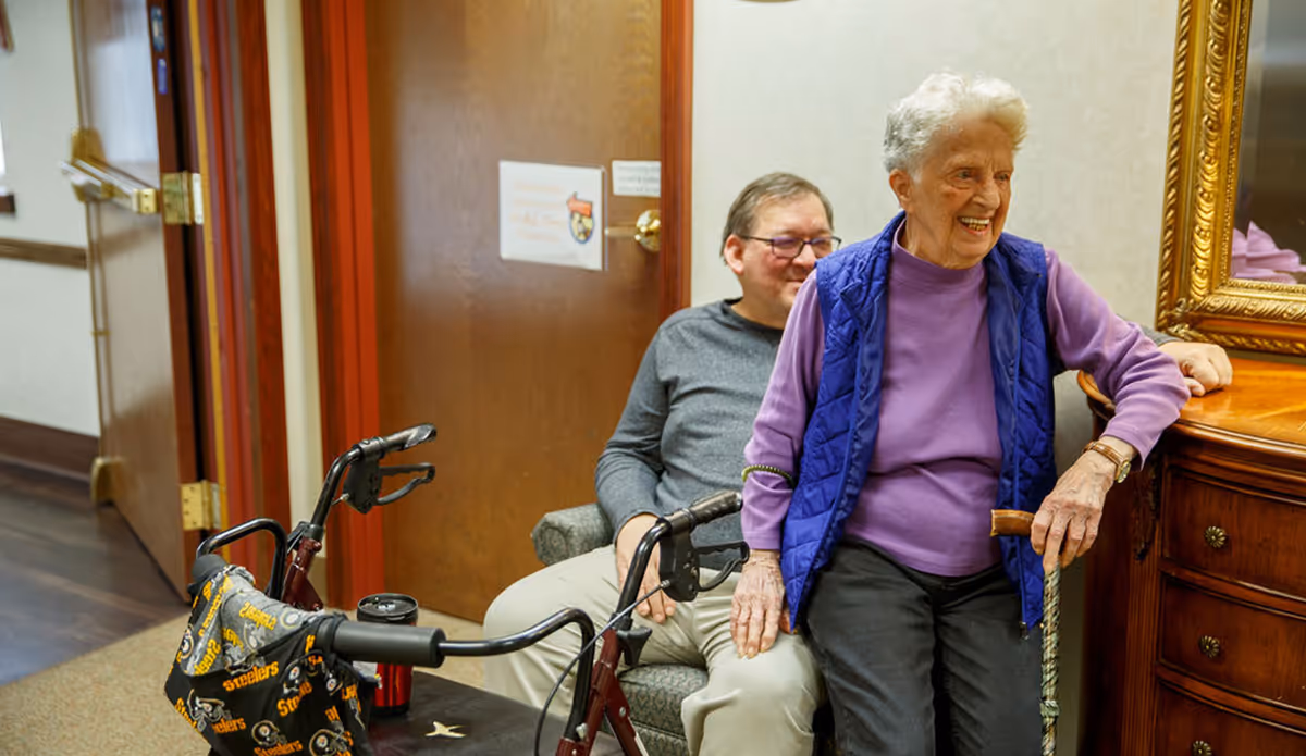 An elderly woman wearing a purple shirt and blue vest is smiling and sitting on a wooden dresser while holding a cane. A man in a gray shirt and glasses is sitting in a chair behind her. A walker with a Pittsburgh Steelers-themed bag is in the foreground. The setting appears to be a hallway in a senior living facility.