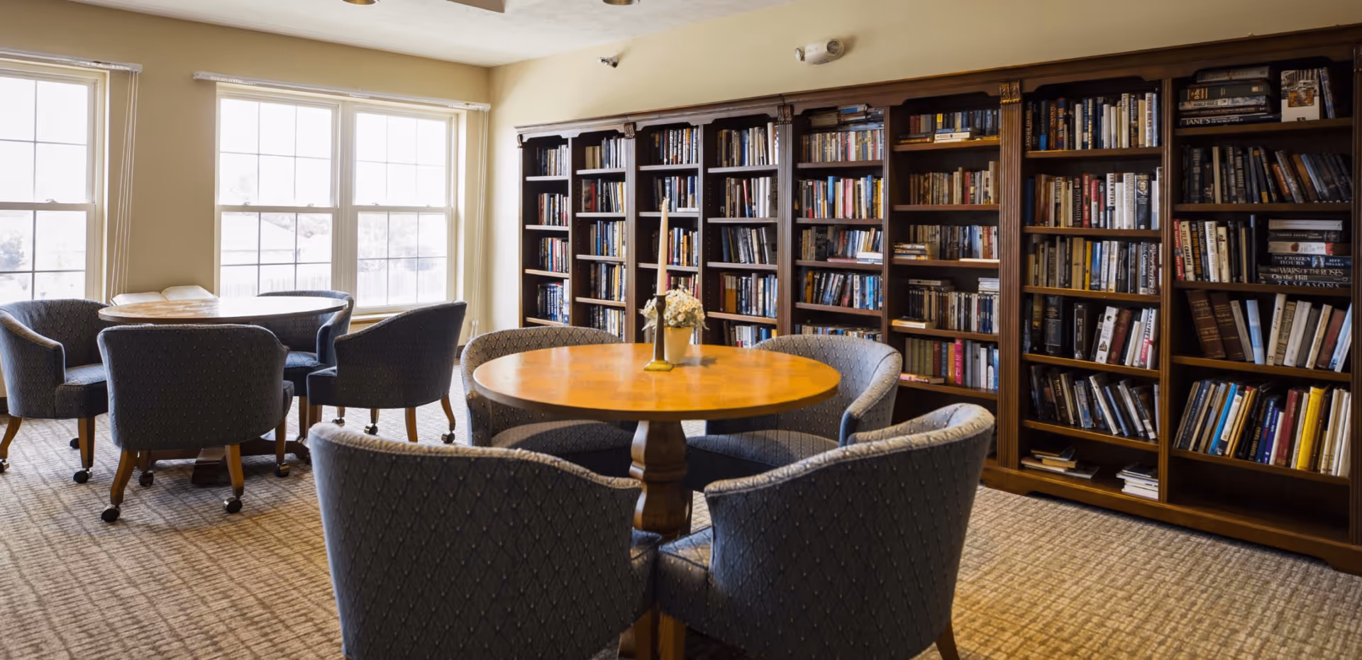 A cozy reading or sitting area in a senior living facility with round wooden tables surrounded by cushioned chairs on wheels. A large wooden bookshelf filled with books lines the wall. Large windows allow natural light to brighten the room.