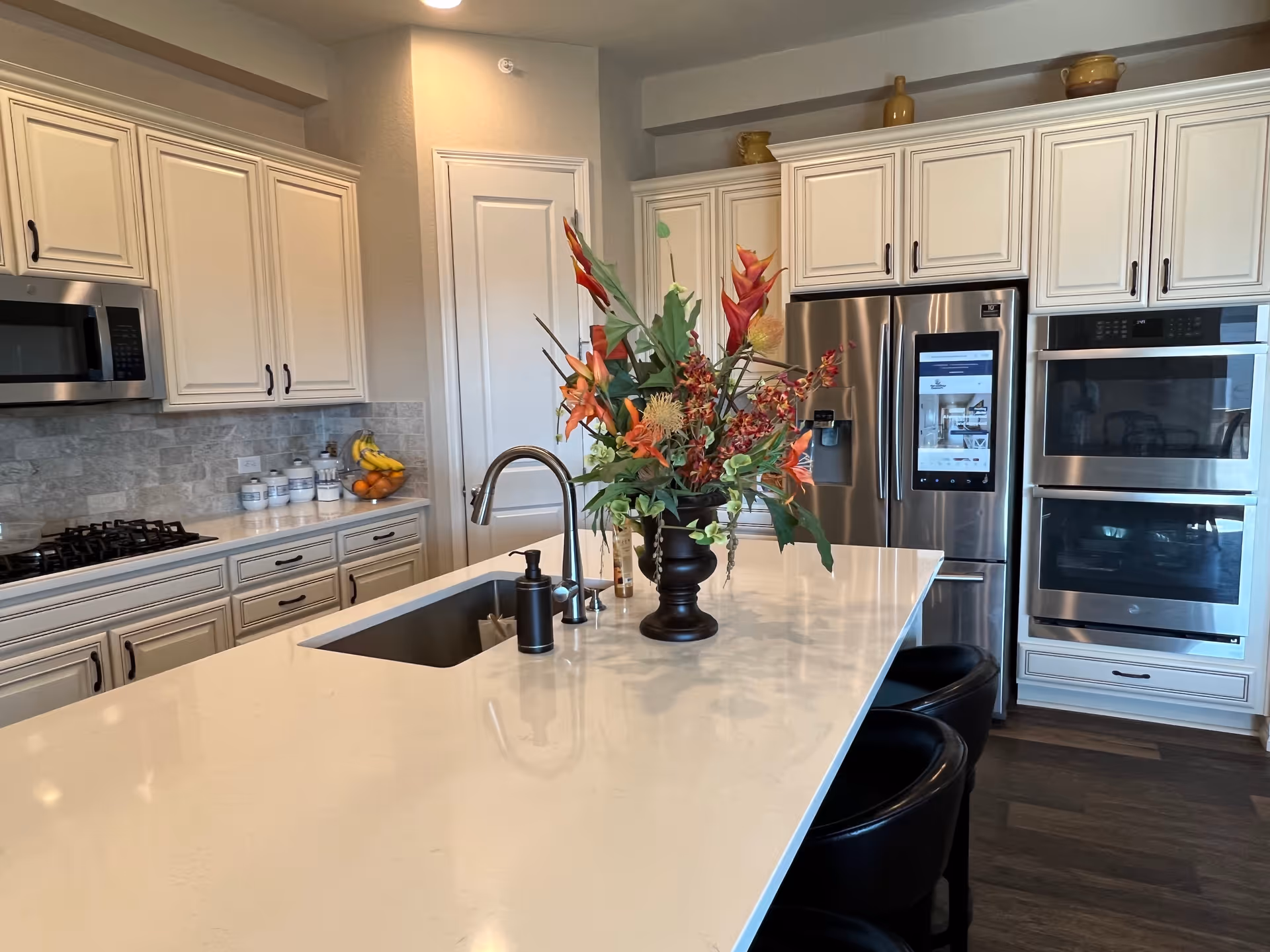 Modern kitchen with white cabinetry, a large white island countertop with a built-in sink and faucet, a black soap dispenser, and a decorative vase with colorful flowers. Stainless steel appliances including a refrigerator with a digital screen, a microwave, and double ovens are visible. The kitchen has a light gray tiled backsplash and dark wood flooring.