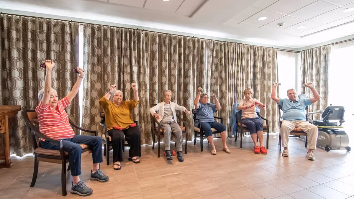 Six elderly individuals seated in chairs in a room with patterned curtains, participating in a group exercise session by lifting small dumbbells above their heads.