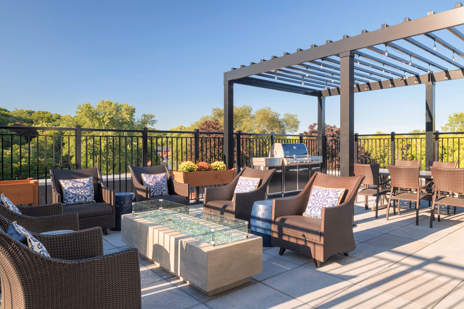 Outdoor patio area with wicker chairs and patterned cushions arranged around a rectangular fire pit table. There is a pergola structure with string lights overhead and a stainless steel grill in the background. The patio is surrounded by a black metal fence and lush green trees are visible beyond.
