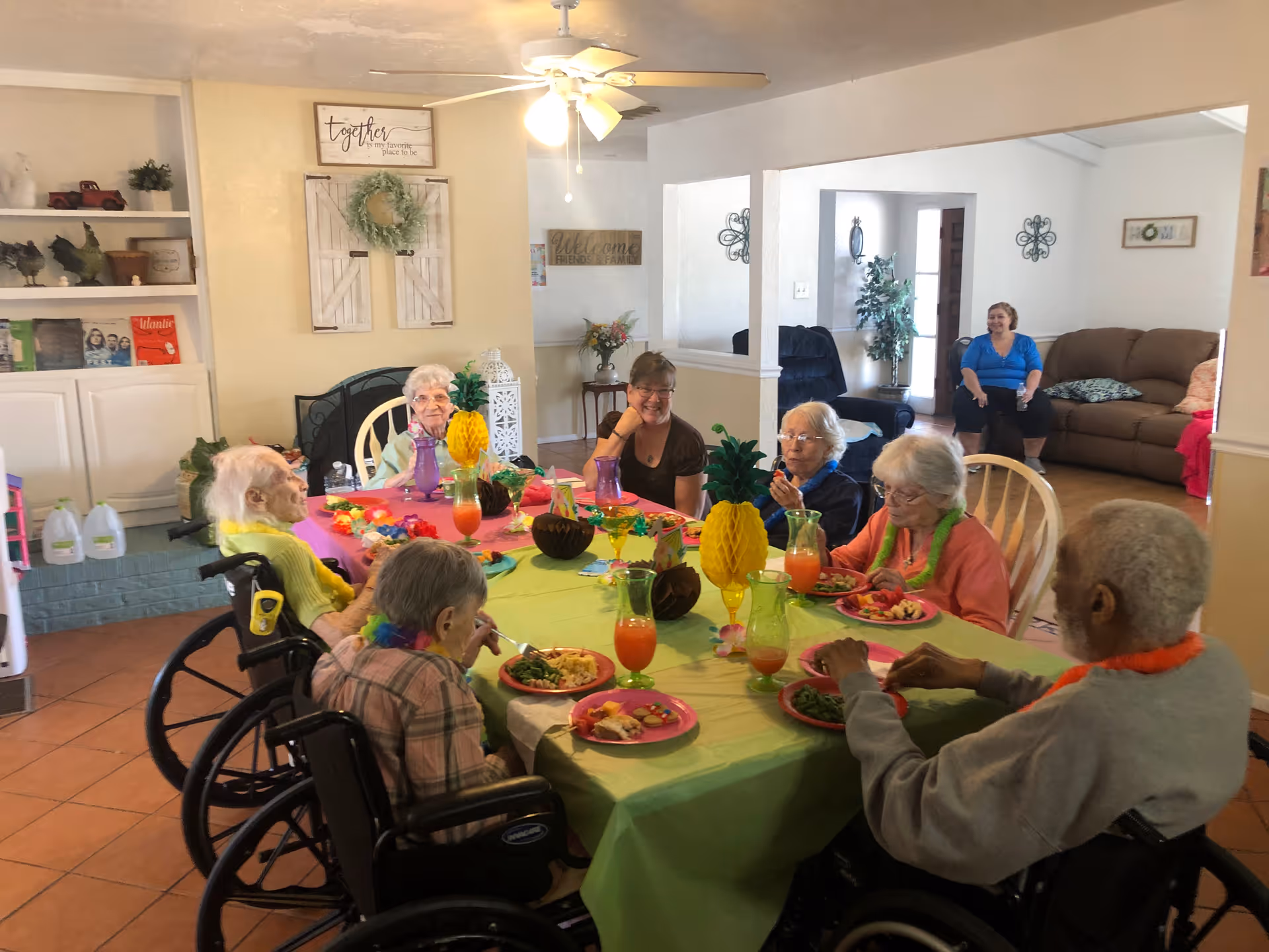Elderly residents and a staff member seated around a brightly decorated table enjoying a meal in a communal dining room of an assisted living facility.