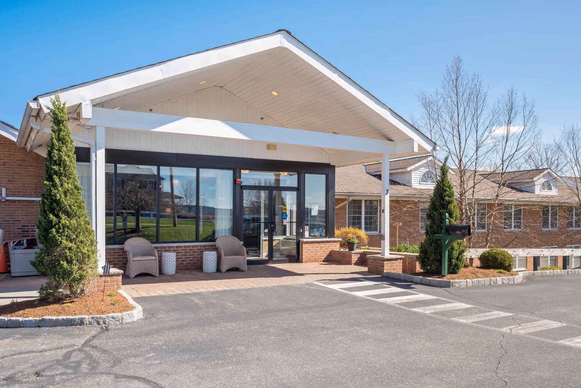 Entrance of Bentley Assisted Living at Branchville showing a covered porch with two wicker chairs and small tables, brick exterior walls, glass doors and windows, landscaped plants, and a clear blue sky.