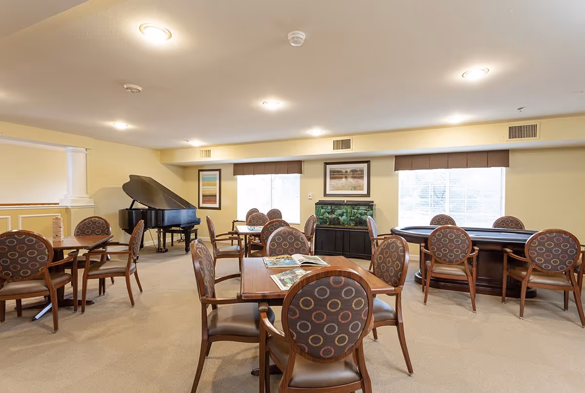 A well-lit common area with several round tables and chairs featuring patterned upholstery. There is a grand piano in the corner, a fish tank against the wall, and framed artwork above it. Large windows with blinds allow natural light into the room.