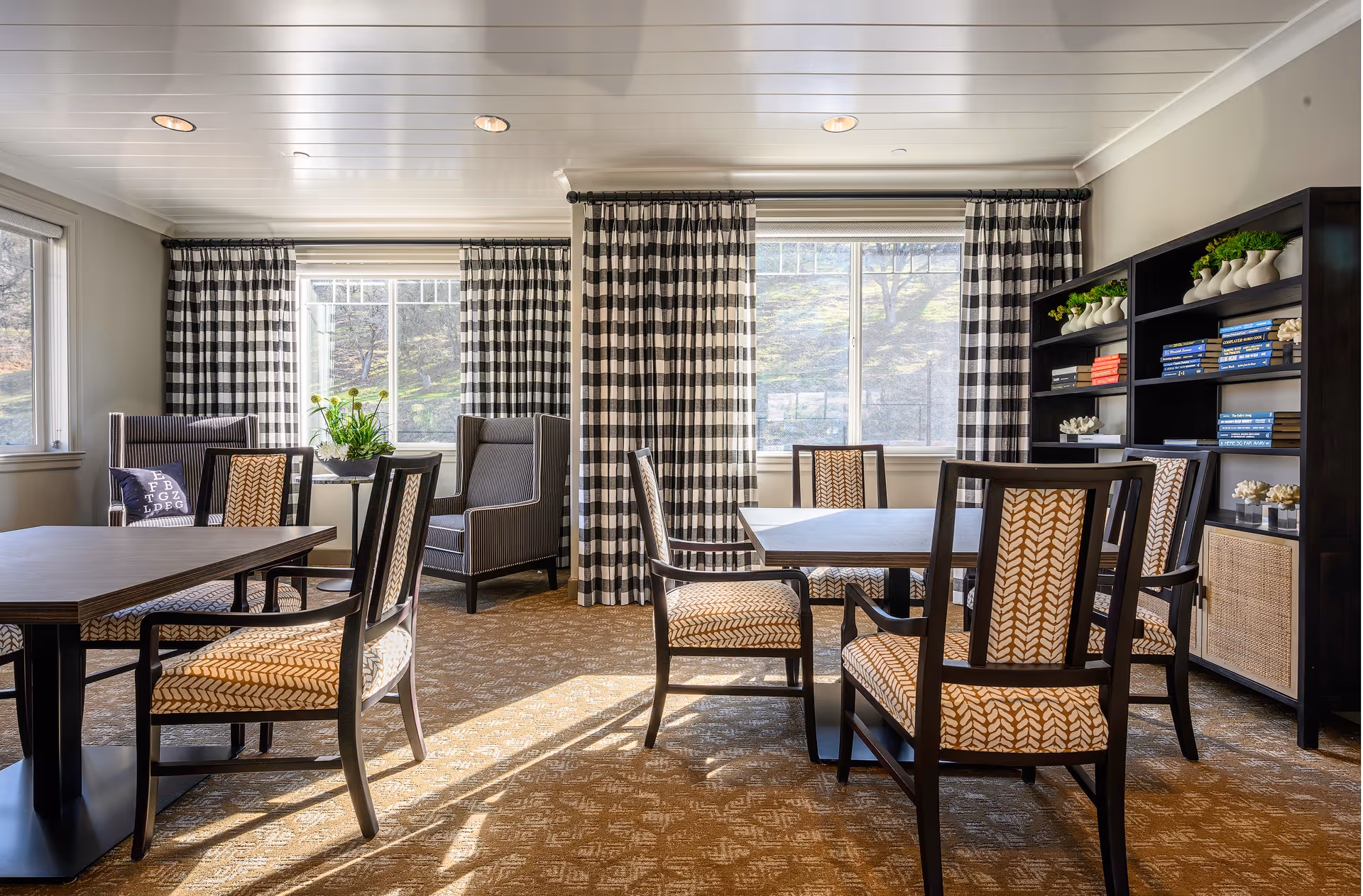 Sunlit communal dining area with patterned chairs around tables, armchairs, bookshelves and large checked curtains.