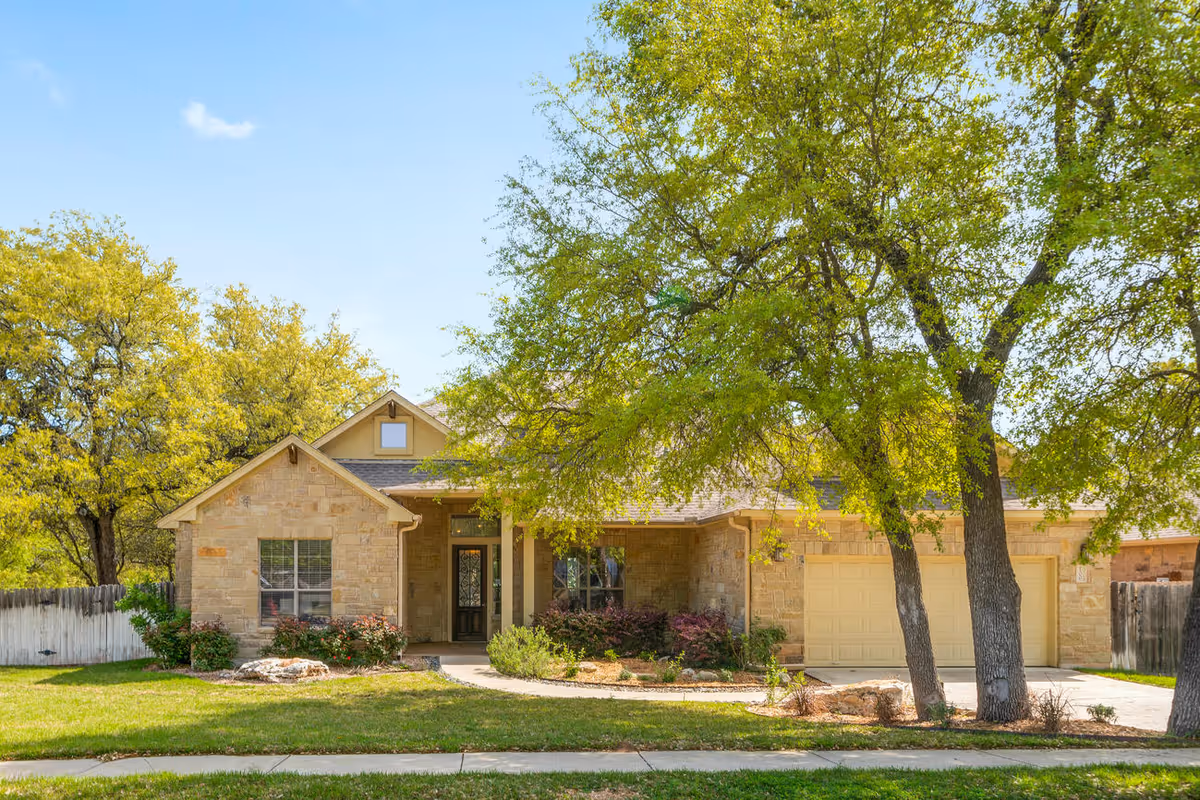 A single-story stone house with a front porch, two large trees in the front yard, a driveway leading to a two-car garage, and a well-maintained lawn with bushes and plants under a clear blue sky.