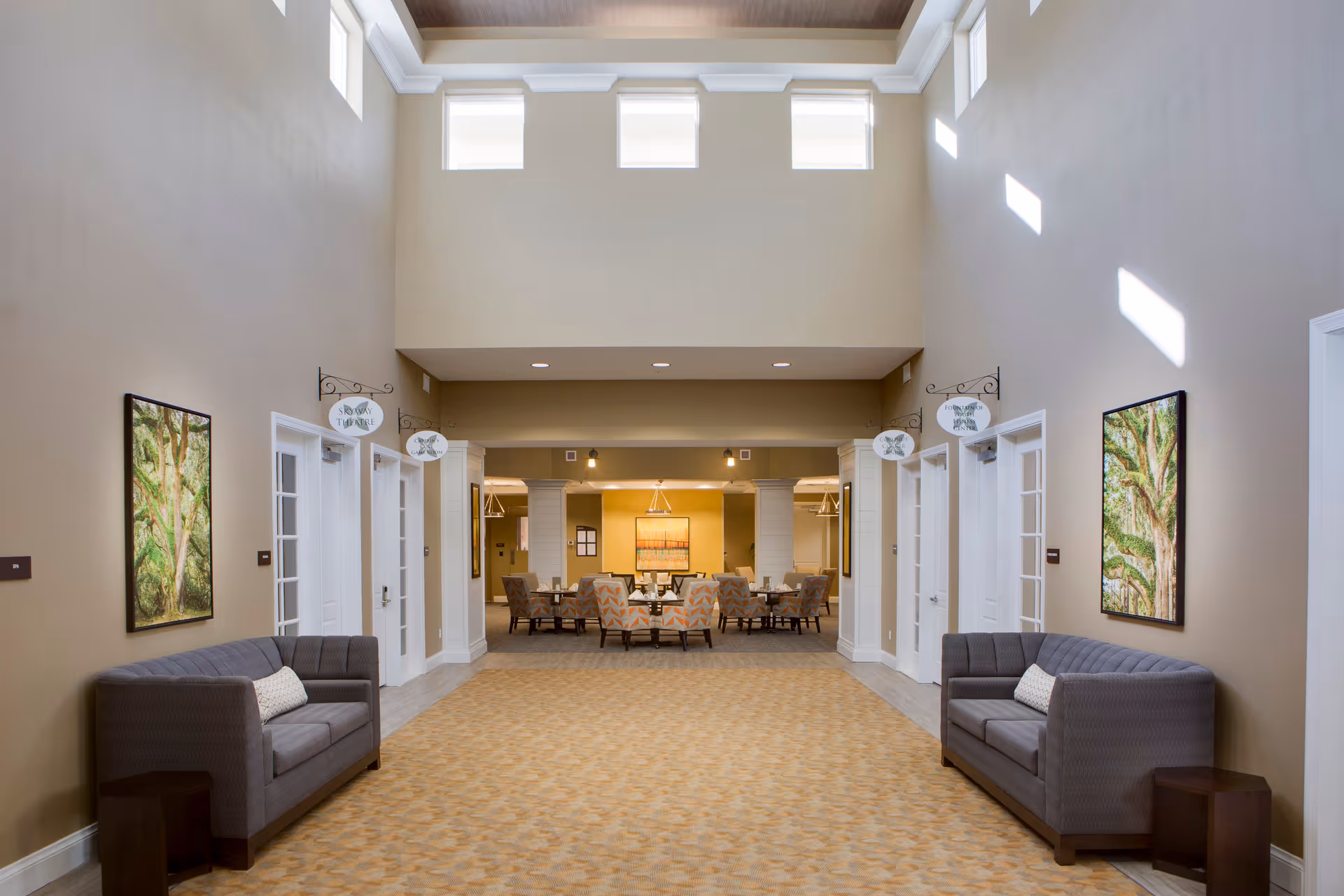 A spacious, well-lit common area with high ceilings and multiple windows near the ceiling. The room features two gray sofas with pillows on either side of the hallway, each accompanied by a small wooden side table. There are framed paintings of trees on both walls. At the end of the hallway, there is a seating area with several upholstered chairs and tables under warm lighting. Signs above the doors on both sides read 'Sun Room' and 'The Office'.