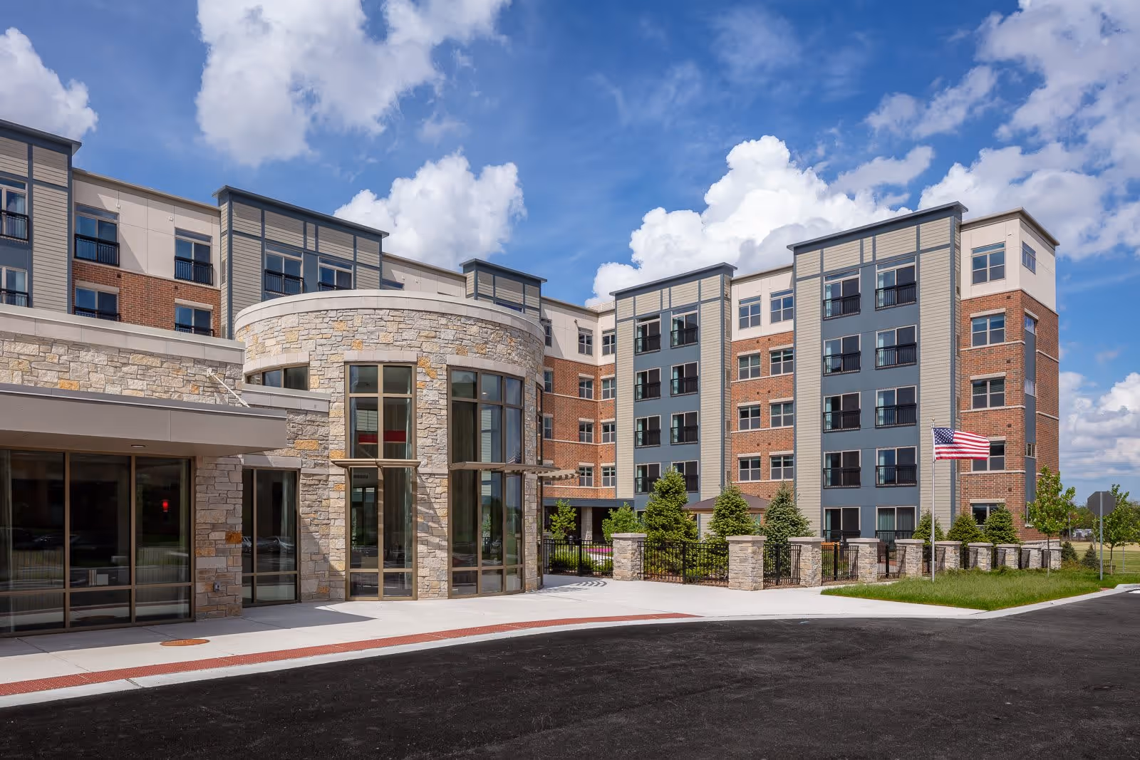 Exterior view of a multi-story senior living facility building with a mix of stone, brick, and siding materials. The entrance features large glass windows and a curved stone facade. There is a paved driveway and a small landscaped area with trees and an American flag on a flagpole. The sky is partly cloudy.