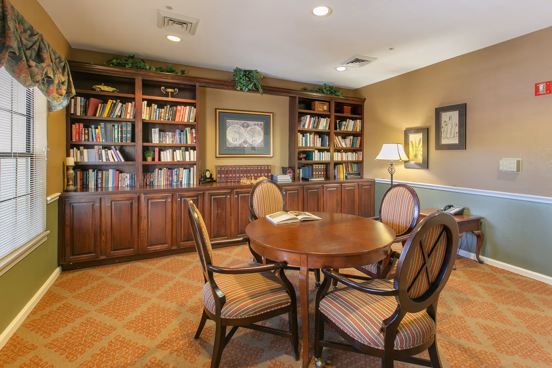 A cozy interior room featuring a round wooden table with four upholstered chairs arranged around it. Behind the table is a large wooden bookshelf filled with books and decorative items. The room has warm lighting, framed artwork on the walls, a window with patterned curtains, and a carpet with a geometric pattern.
