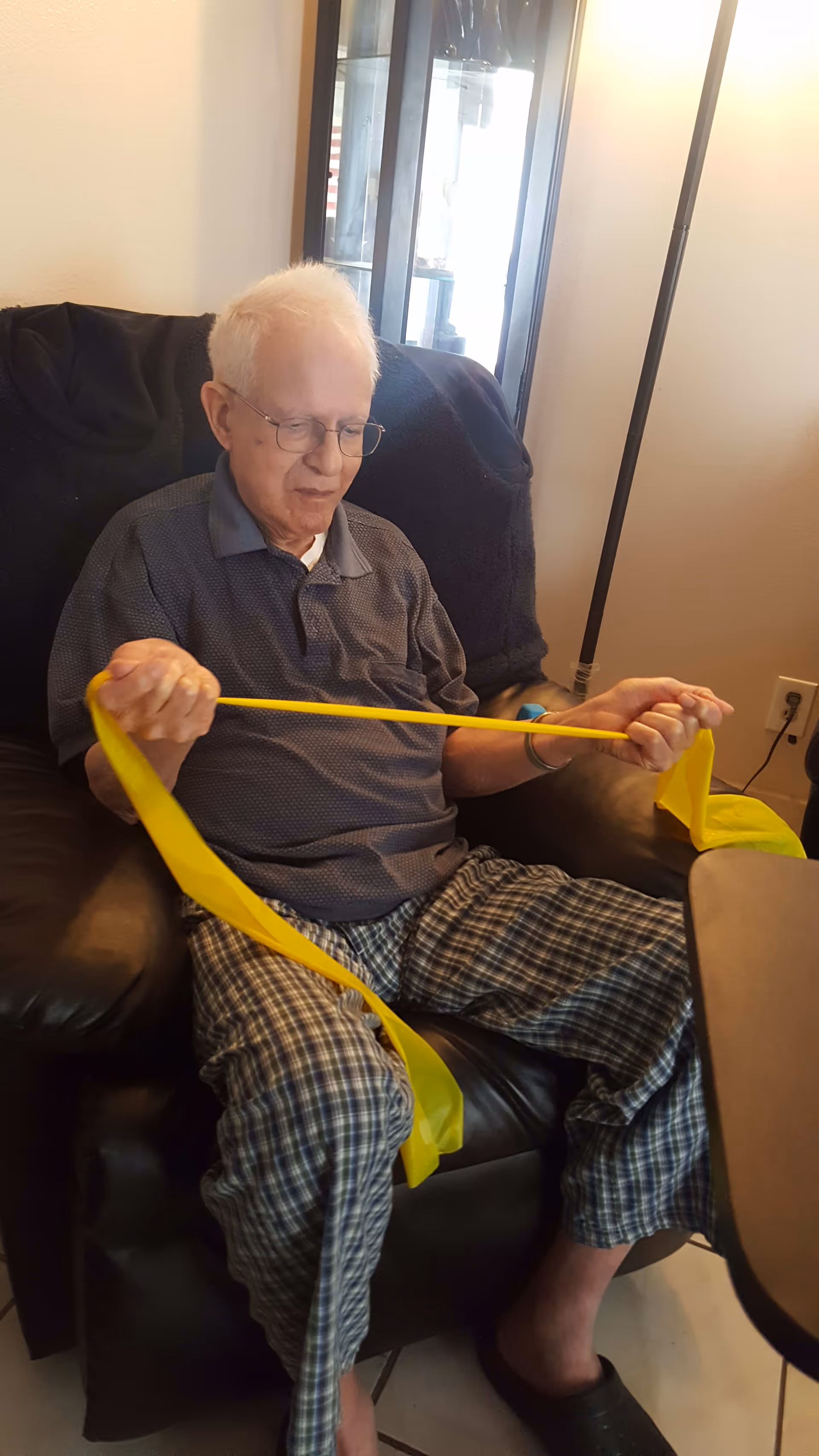 An elderly man with white hair and glasses is sitting in a black recliner chair indoors, exercising with a yellow resistance band. He is wearing a dark polo shirt and plaid pajama pants. Behind him is a glass cabinet and a floor lamp is visible to the right.