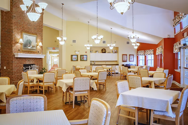 Bright, spacious dining room with many cloth-covered tables and upholstered chairs, chandeliers, a brick fireplace and large windows.
