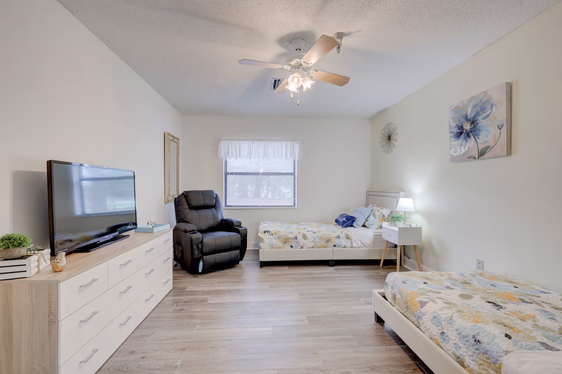 A bright and clean bedroom with two single beds covered in floral-patterned quilts, a black recliner chair near a window with white curtains, a white dresser with a flat-screen TV on top, and a small white nightstand with a lamp and plant. The walls are light-colored with a floral painting and a decorative clock, and the floor is wood-style tile.