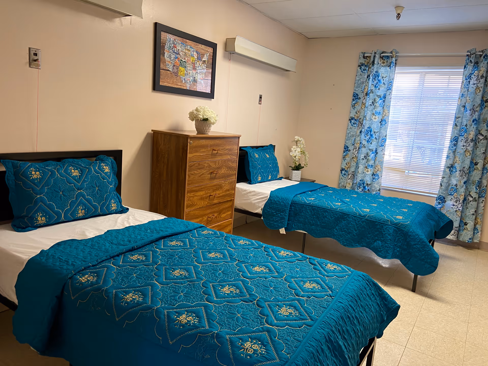 Two twin beds with turquoise quilts in a bright shared bedroom, dresser, and blue curtains.