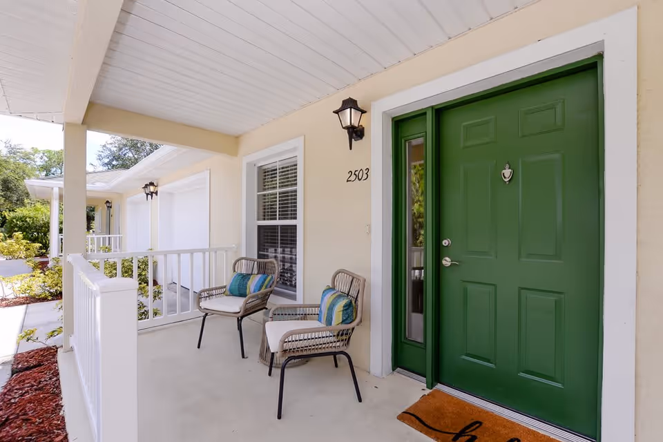 Front porch of a residential unit with a green door numbered 2503, two wicker chairs with cushions, a welcome mat, white railing, and exterior wall lights under a covered roof.