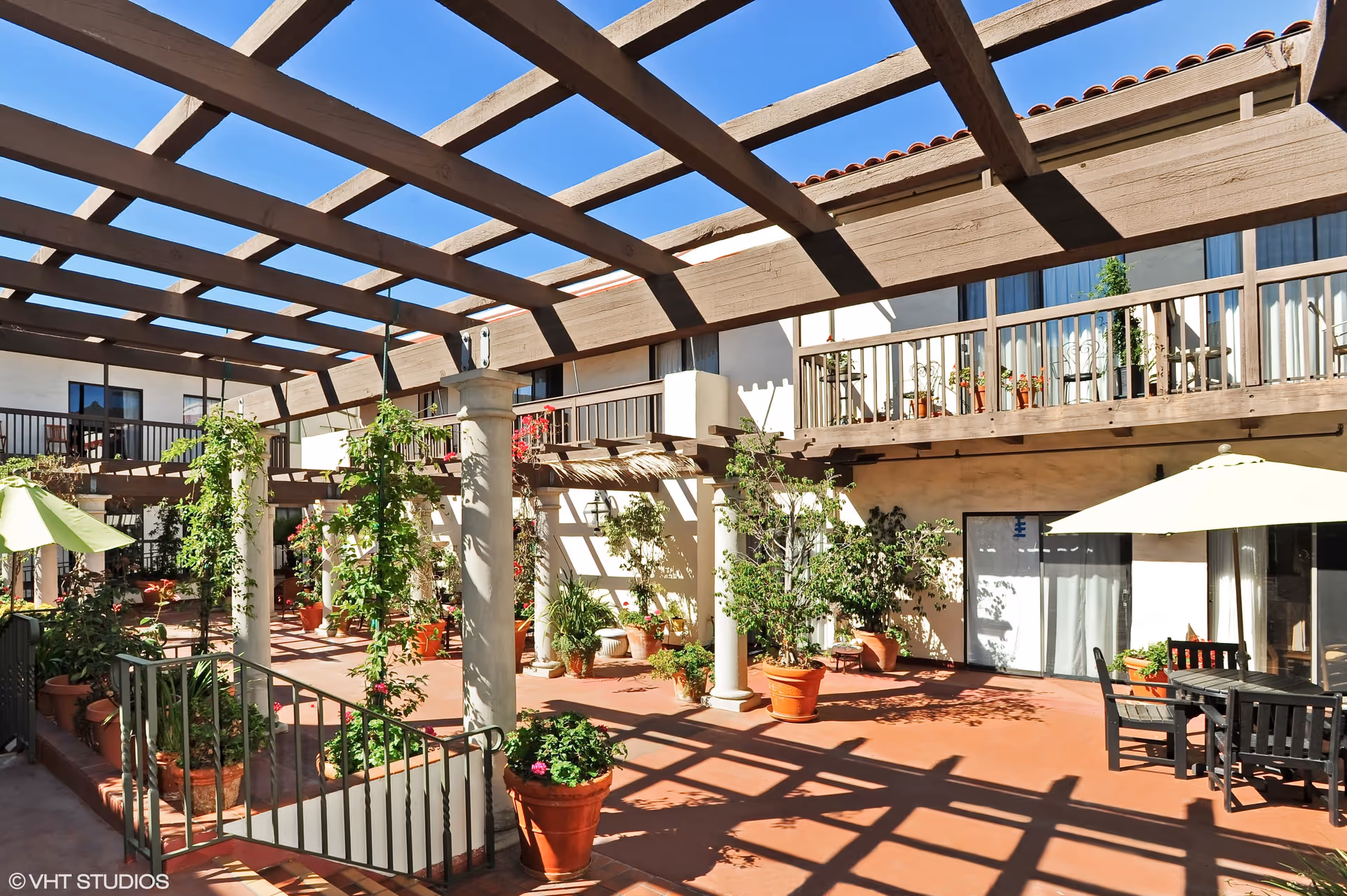 Outdoor courtyard area with a wooden pergola casting shadows on the ground. There are several potted plants and climbing vines around the space. Patio furniture with umbrellas is visible, and the courtyard is surrounded by a two-story building with balconies and sliding glass doors.