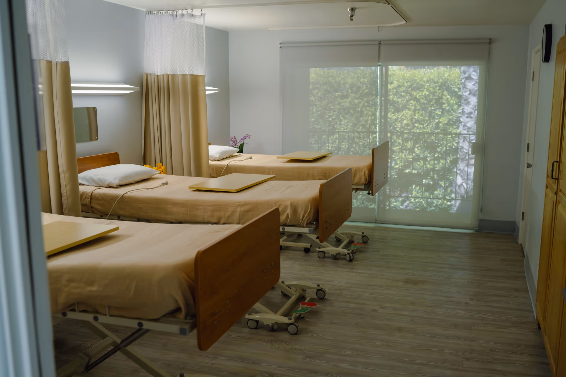 A hospital room with three beds arranged in a row, each bed covered with beige sheets and equipped with a pillow and a movable overbed table. The room has light-colored walls, wooden flooring, and large windows with blinds that let in natural light. Privacy curtains hang between the beds.