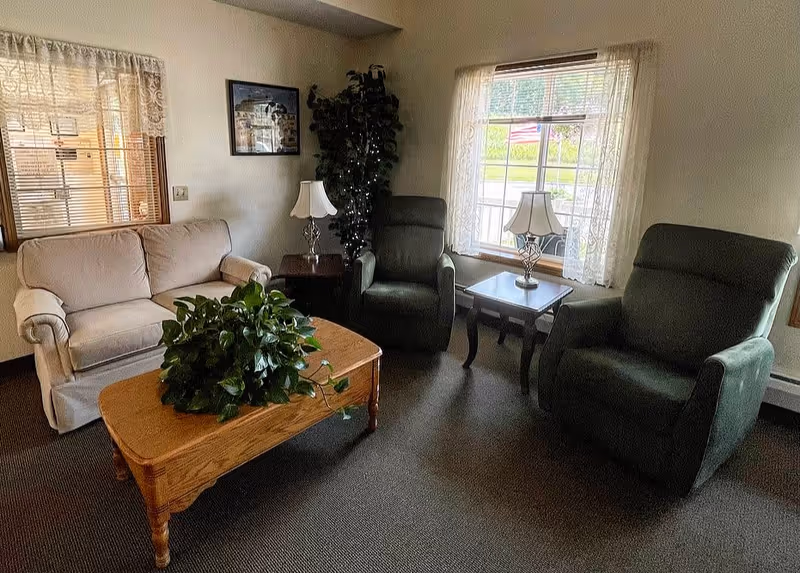 Small living room with a loveseat, two recliners, a wooden coffee table topped with a plant, and side tables with lamps by windows.