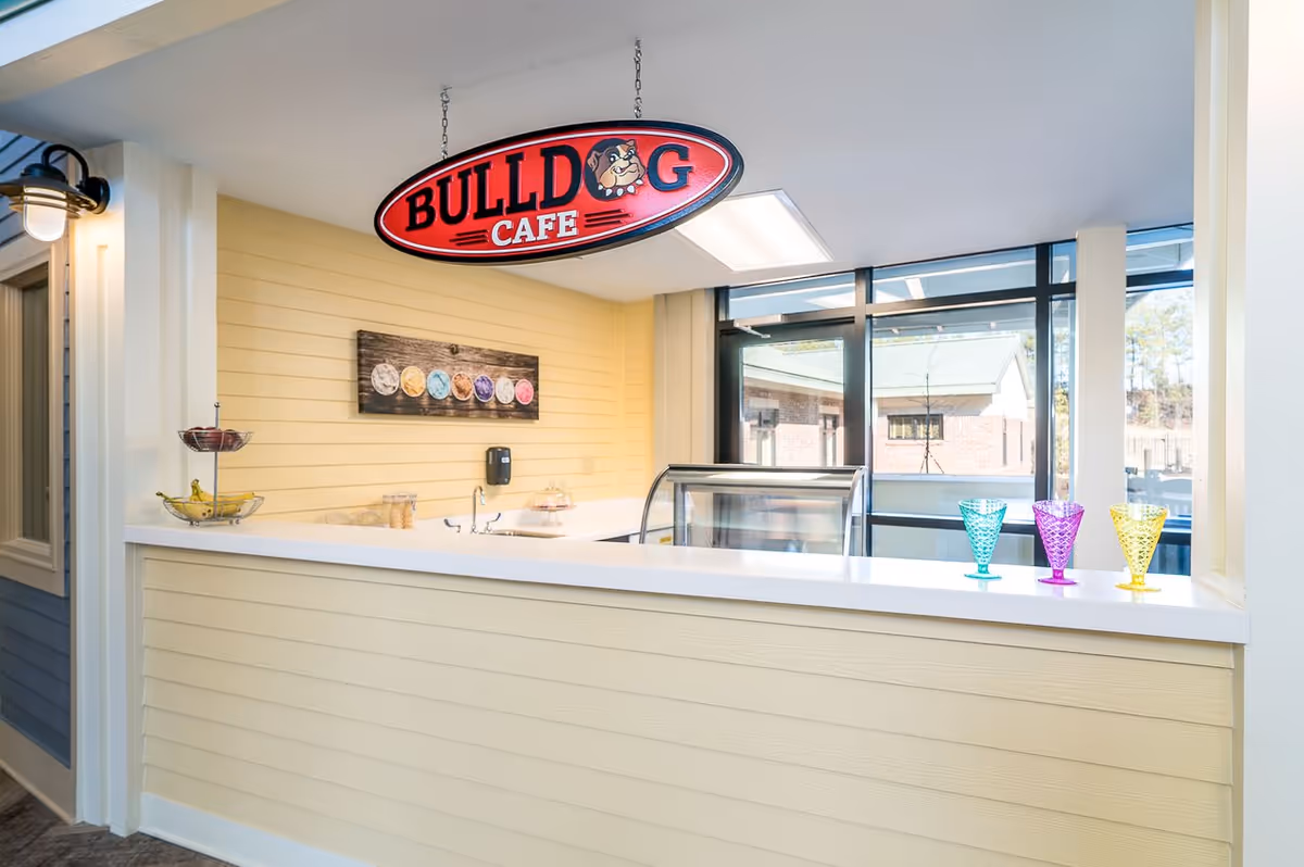 Interior view of a small cafe counter with a hanging sign that reads 'BULLDOG CAFE'. The counter has a white surface with a tiered fruit basket holding bananas and apples, a display case, and three colorful glass cups in blue, purple, and yellow. The walls are painted light yellow with a decorative picture of ice cream scoops. Large windows in the background show an outdoor view of buildings and trees.