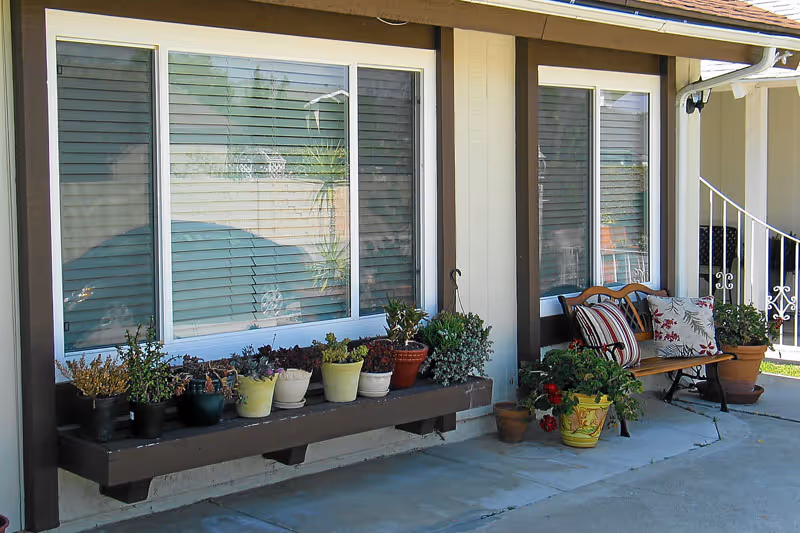 Outdoor patio area of a facility with a row of potted plants on a wooden shelf attached to the wall beneath two large windows. To the right, there is a wooden bench with decorative cushions and additional potted plants on the ground. The area is paved with concrete and part of a white railing is visible in the background.