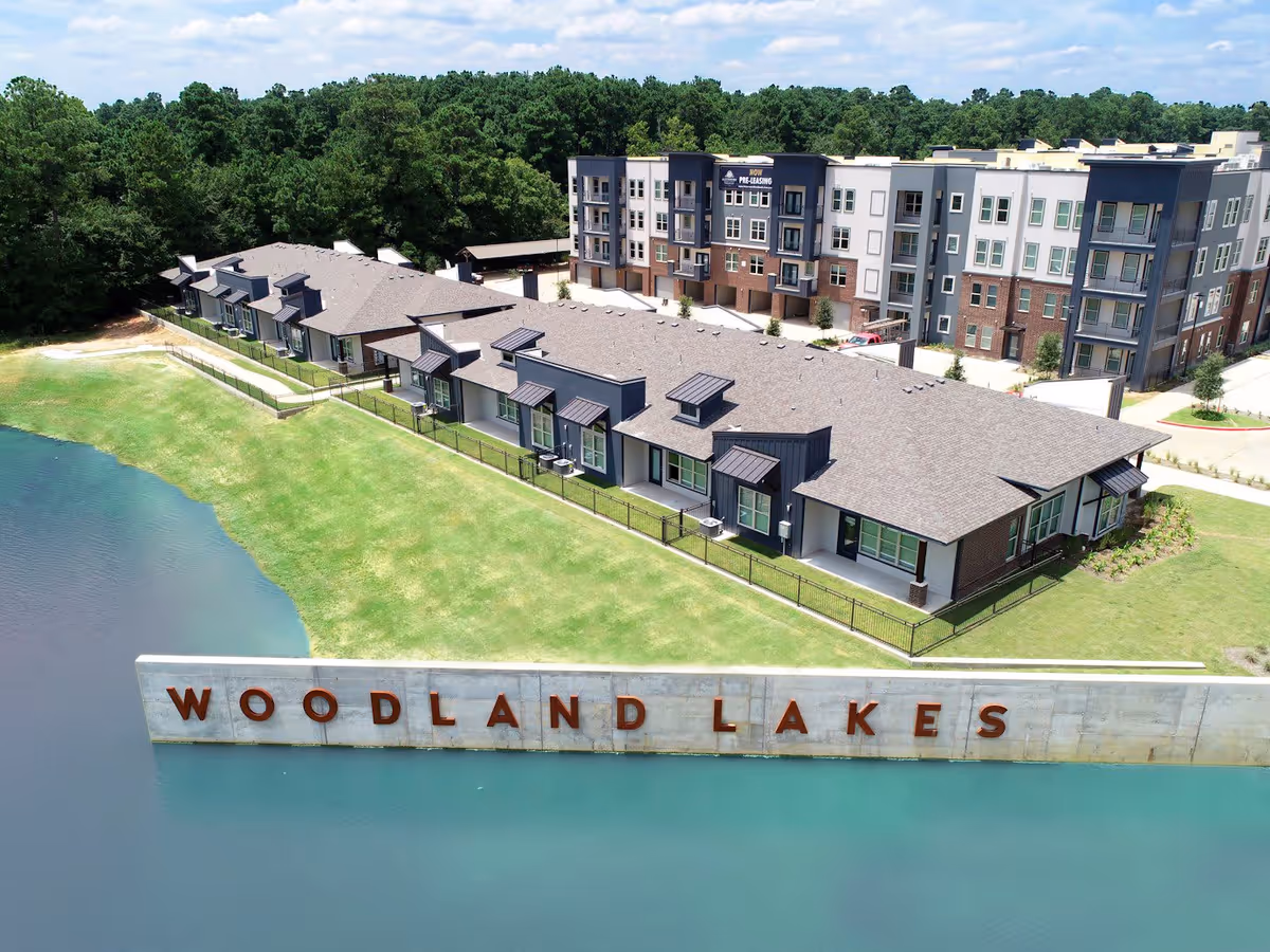 Aerial view of a senior living facility named Woodland Lakes featuring a multi-story building and a row of single-story units next to a body of water with a concrete wall displaying the name 'WOODLAND LAKES'. The area is surrounded by green grass and trees under a partly cloudy sky.
