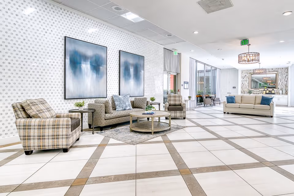 A spacious and bright living room area with patterned tile flooring and white walls featuring a subtle polka dot design. The room is furnished with a beige sofa with blue pillows, two plaid armchairs, a patterned loveseat, and a round wooden coffee table on a small rug. Two large abstract paintings hang on the wall above the loveseat. In the background, there is a large mirror above a white cabinet and two modern chandeliers hanging from the ceiling. Large windows and glass doors allow natural light to enter the room.