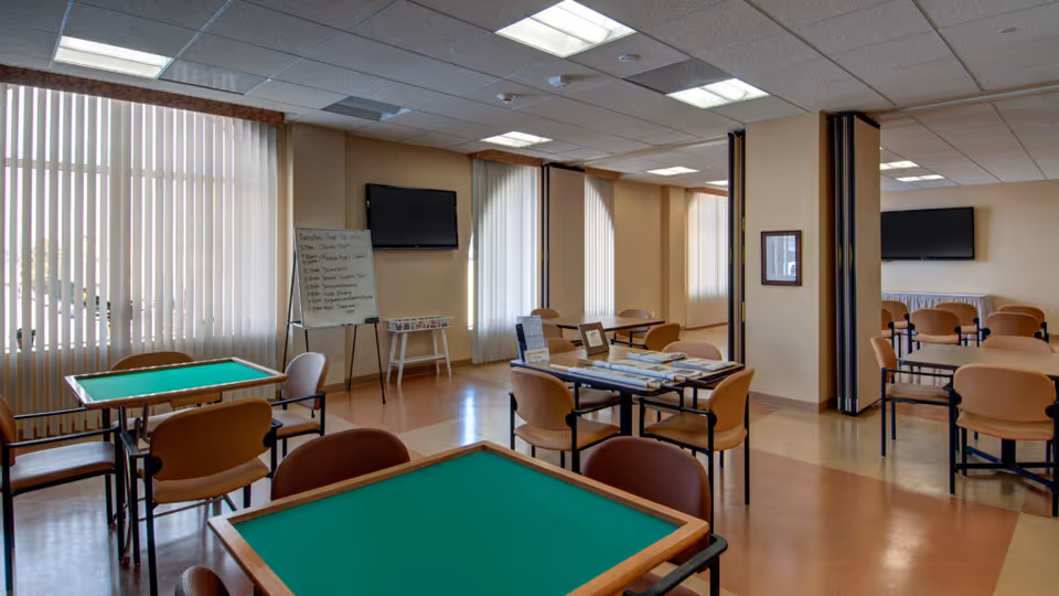 Well-lit communal activity room with multiple card tables and chairs, wall-mounted TVs, and large windows with vertical blinds.