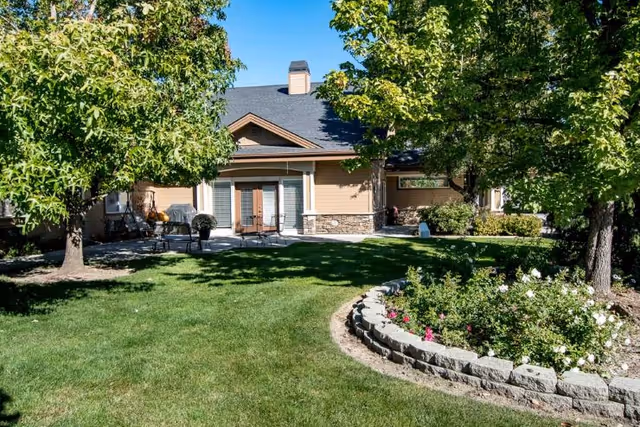 Sunny courtyard with a lawn, trees, a flower bed, and the exterior of a single-story memory care building with a small patio.