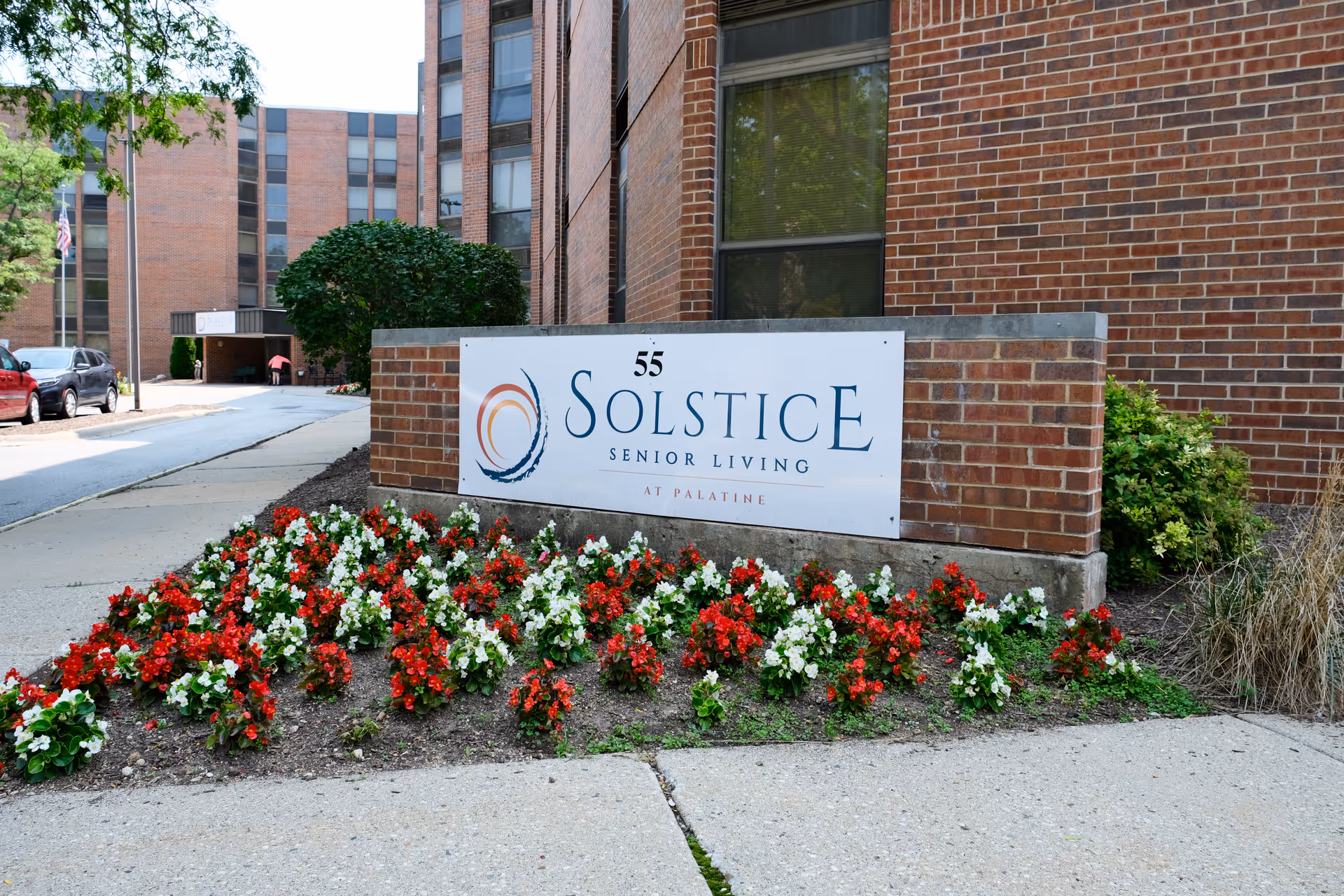Brick building entrance with a 'Solstice Senior Living' sign and a bed of red and white flowers in front.