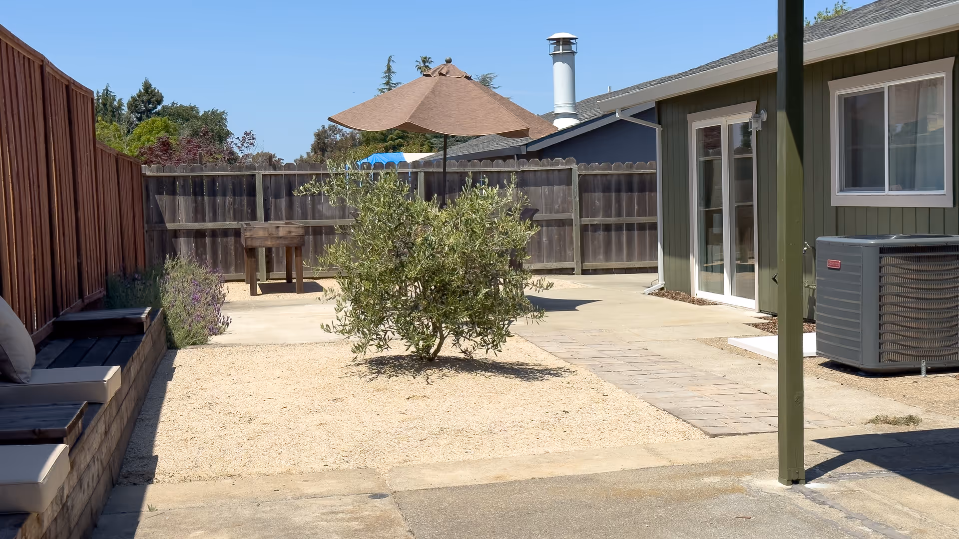 Outdoor patio area with a small tree in the center, surrounded by gravel and concrete. There is a wooden fence in the background, a table with a large umbrella, and a green building with glass doors and windows. A bench with cushions is visible on the left side.