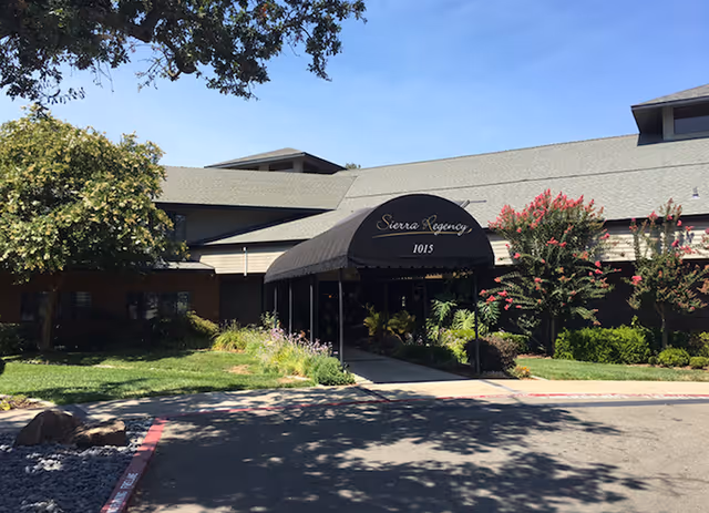 Front exterior view of Sierra Regency facility with a covered entrance canopy displaying the name and address number 1015. The building is surrounded by green landscaping, trees, and flowering bushes under a clear blue sky.